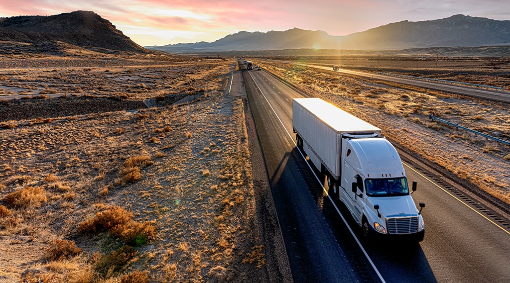 White semi-trailer truck heading down a four-lane highway at dusk.